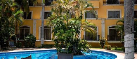 A hotel pool area with a circular pool, palm trees, and a yellow building with balconies.