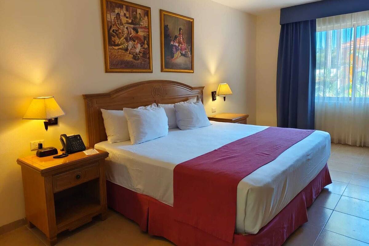 Habitación de hotel con una cama grande, dos lámparas de noche, una mesita de noche de madera con un teléfono y dos cuadros en la pared.