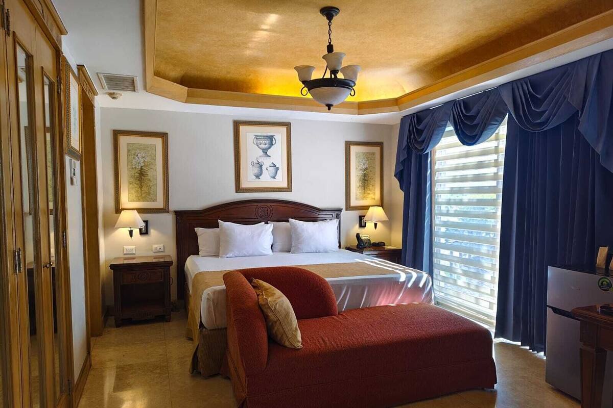 Una habitación de hotel con una cama grande, un sofá rojo, un cabecero de madera, dos mesitas de noche con lámparas y un ventanal con cortinas.