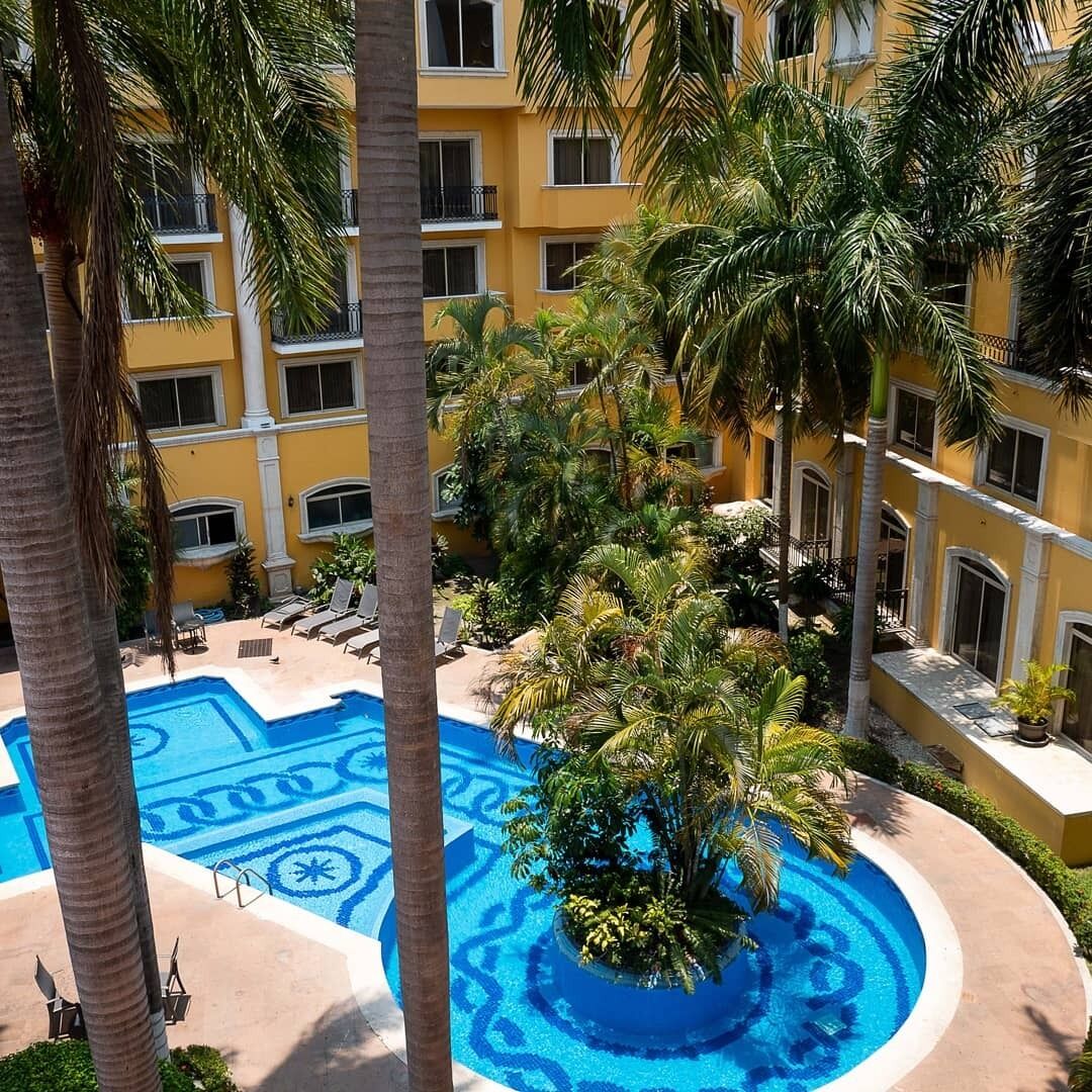 A pool with a blue geometric design surrounded by palm trees and a yellow building with balconies.