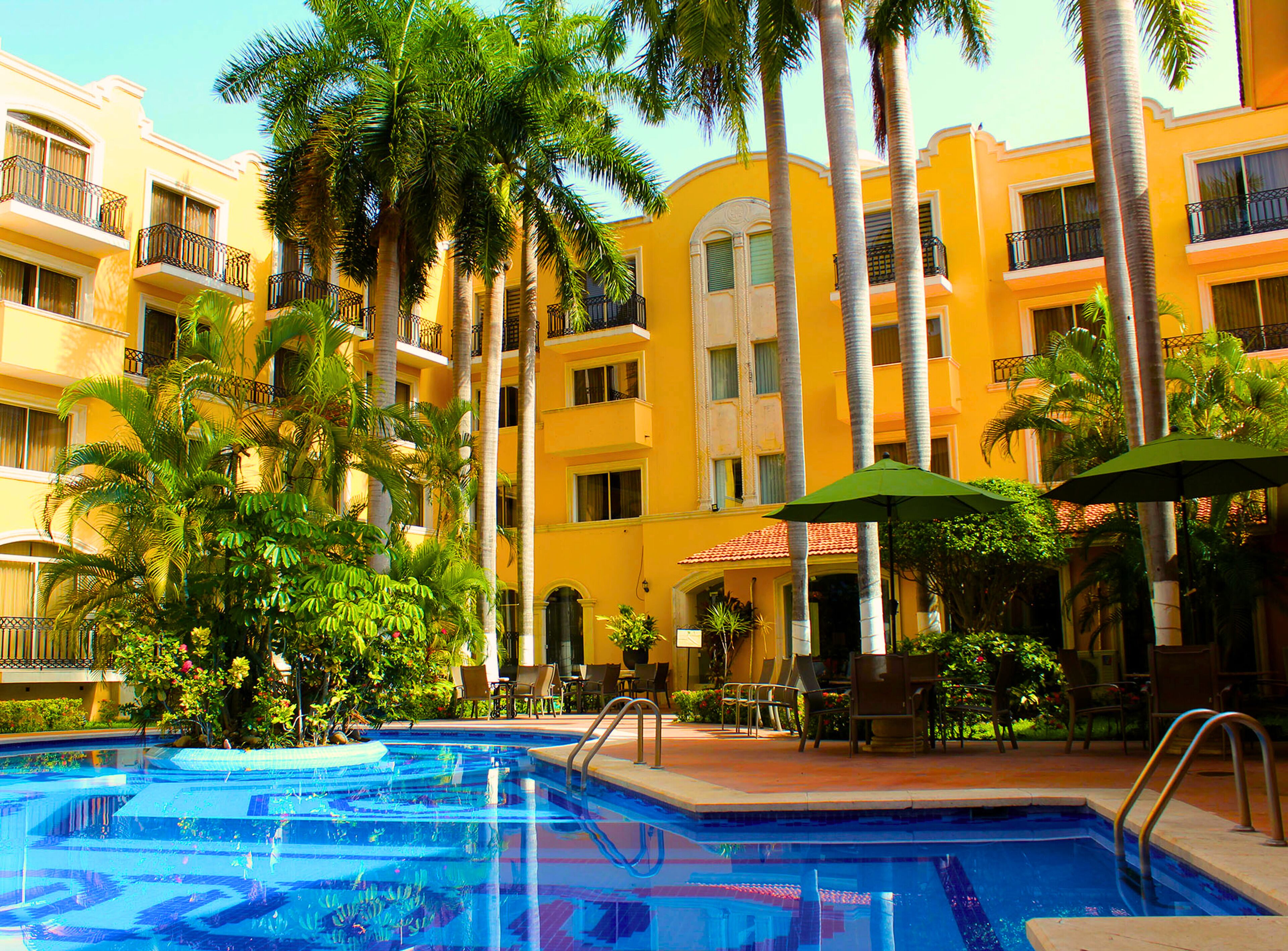 A pool area with a clear blue pool, surrounded by palm trees and yellow buildings with balconies.