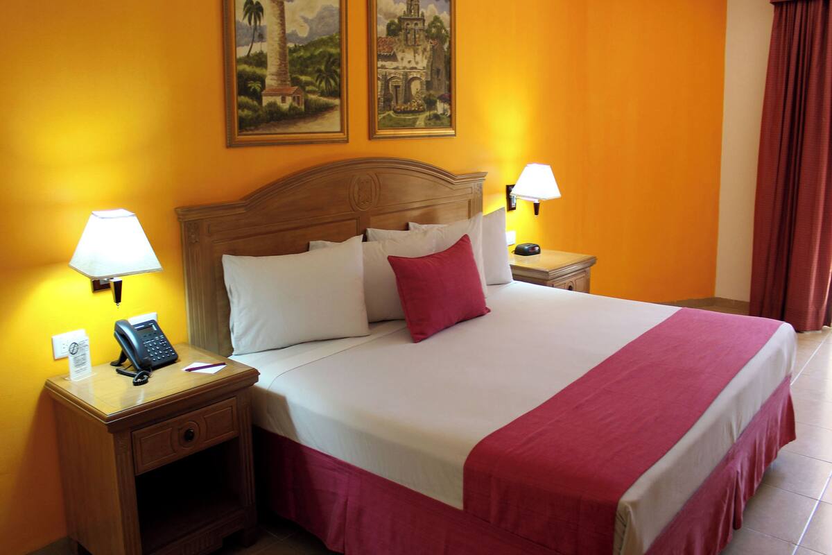 Habitación de hotel con cabecera de madera, cama con ropa de cama blanca y roja, dos lámparas de noche, mesita de noche con teléfono y dos cuadros en la pared.