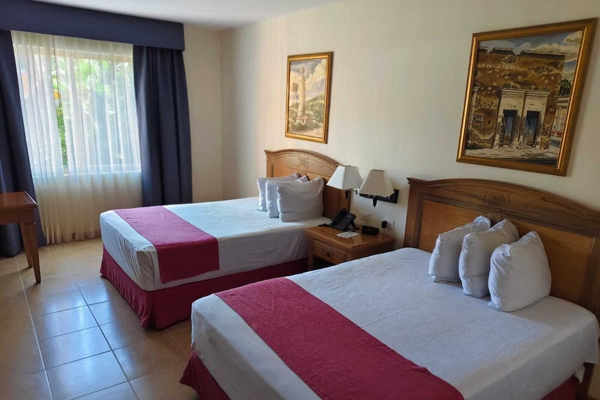 Habitación de hotel con dos camas, una mesita de noche, un escritorio y dos cuadros en la pared.