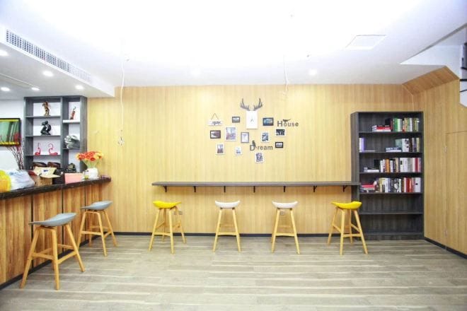 Un área de recepción con mostrador de madera, tres taburetes, una estantería y una pared con cuadros enmarcados y la inscripción “House Dream”.