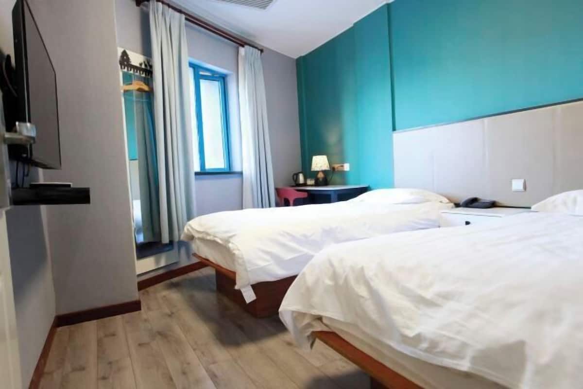 A hotel room with two beds, a television, a window with curtains, and a nightstand with a lamp.
