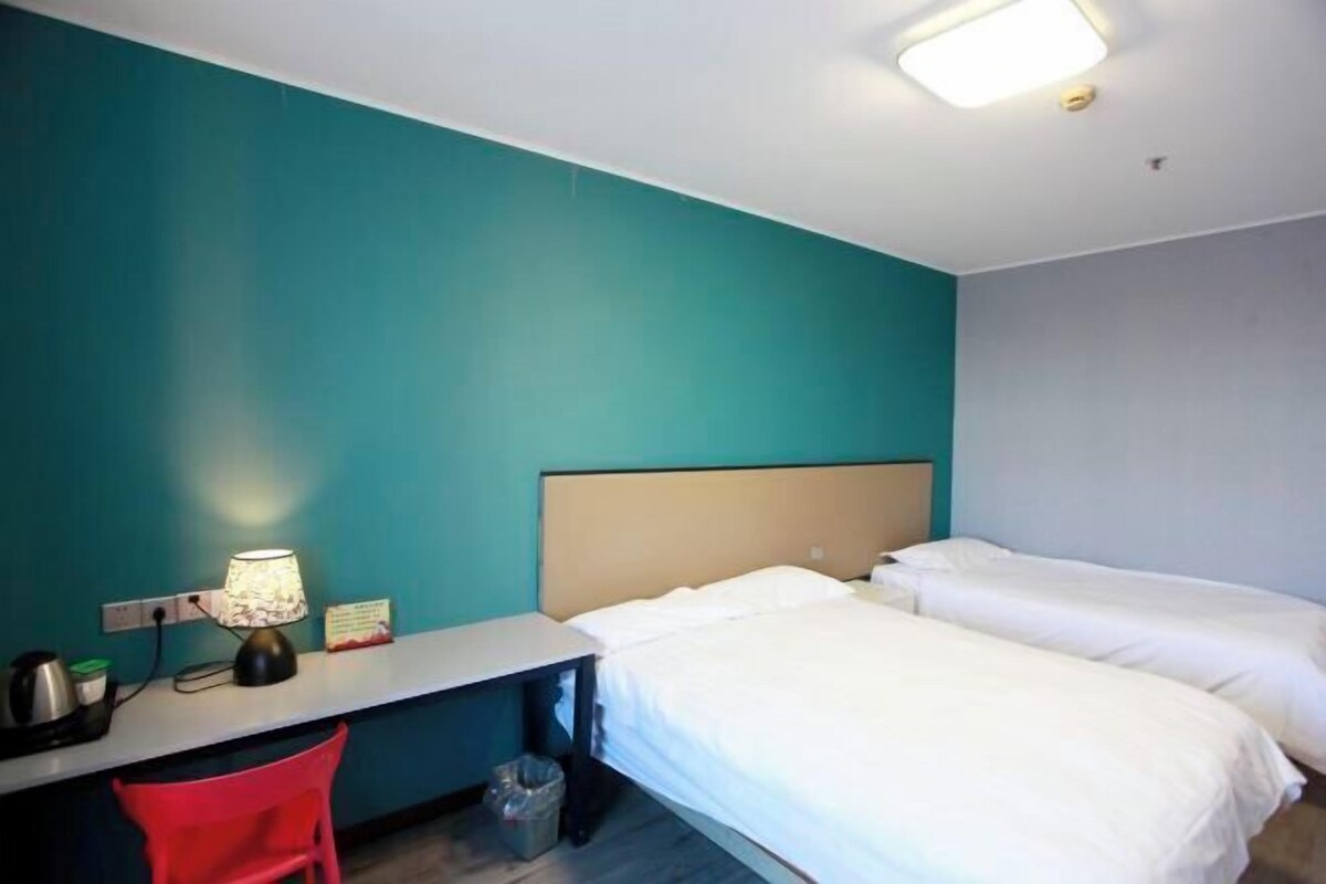 A hotel room with a bed, a desk with a lamp, a chair, and a trash bin.