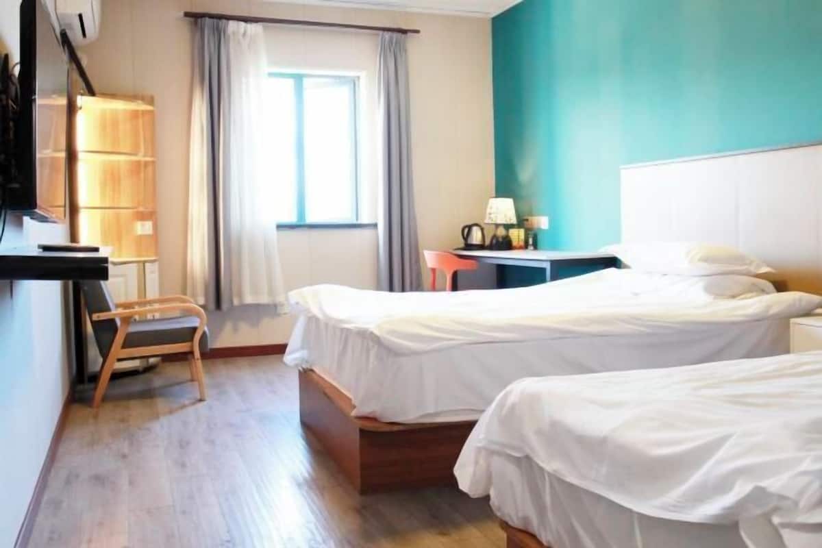 A hotel room with two beds, a desk with a lamp, a chair, a wardrobe, and a window with curtains.