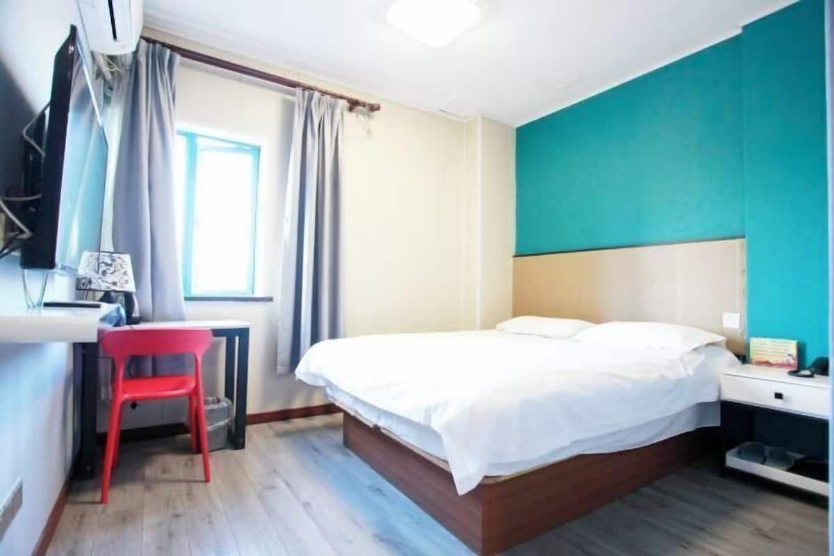 A bedroom with a bed, a desk, a chair, a window with curtains, and a wall-mounted air conditioner.
