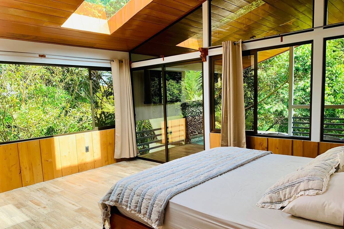 A bedroom with a large bed, wooden walls, and large windows overlooking a garden.