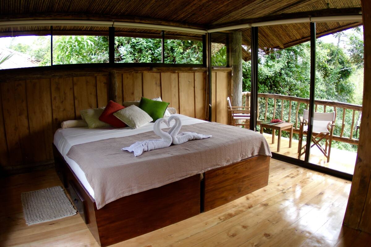 A wooden room with a bed, pillows, and a view of greenery outside.
