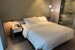 A modern hotel room with a large bed, a bedside table with a lamp, a nightstand, and a glass shower enclosure. - Orange Hotel (Shanghai Jiaotong University, Jiangchuan Road Metro Station) (Shanghai)