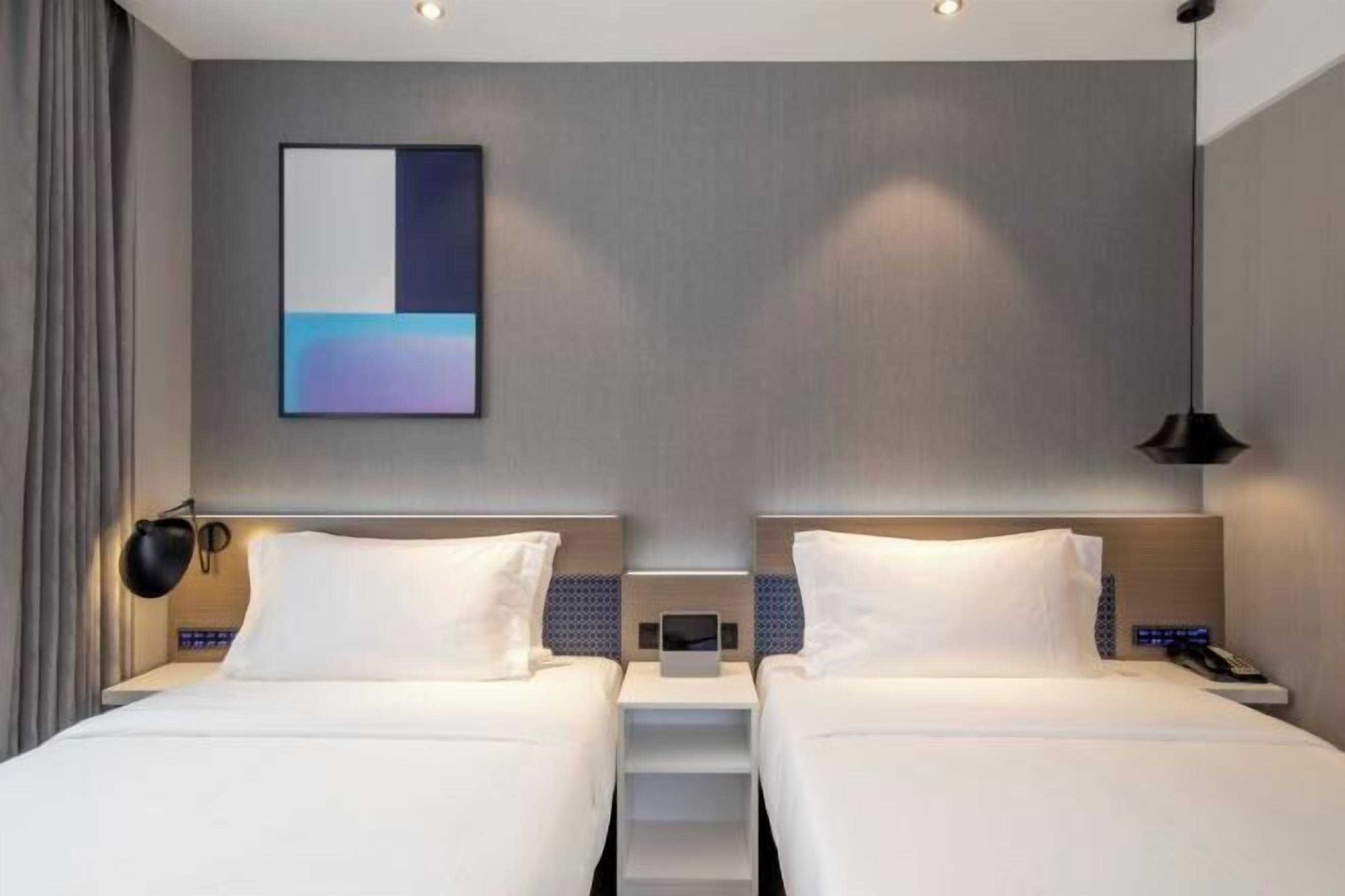 A hotel room with two beds, a bedside table with a lamp, a wall-mounted artwork, and a ceiling light.