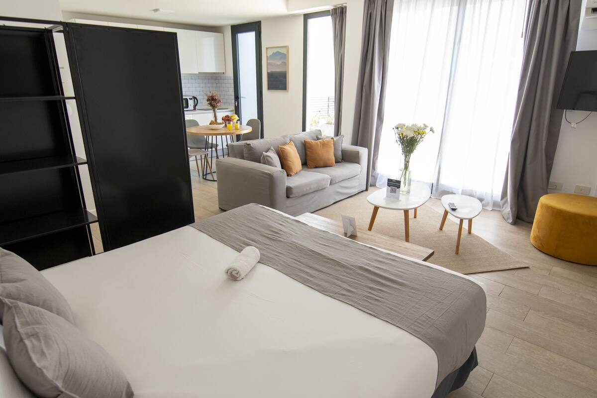 A hotel room with a bed, a sofa, a dining table, and a small table with flowers.