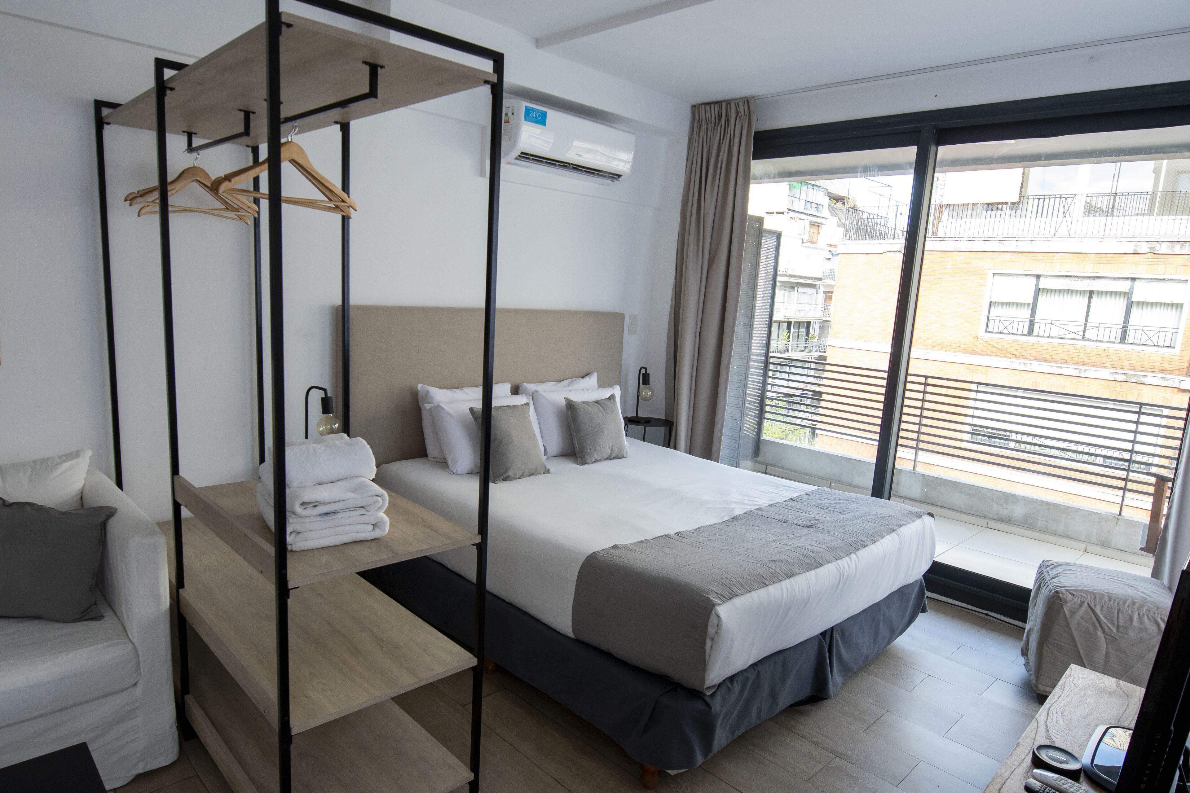 Una habitación de hotel moderna con una cama grande, un área de estar con sofá, vista al balcón y un armario con ropa colgada.