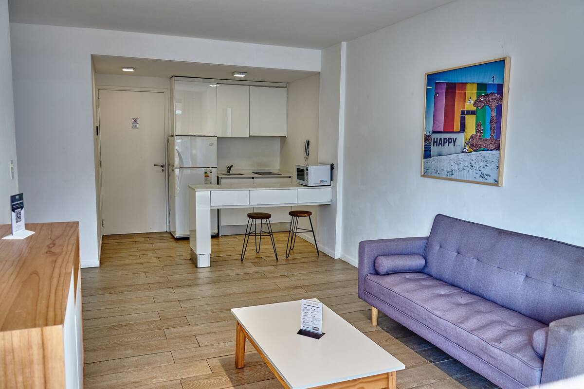 A modern living room with a purple sofa, a wooden coffee table, and a kitchen area with white cabinets and a microwave.