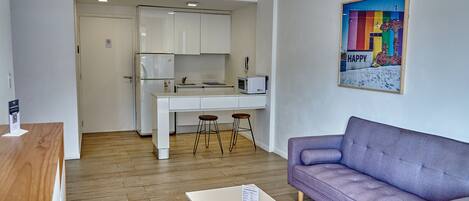 Una sala de estar moderna con un sofá morado, una mesa de centro de madera y una cocina con armarios blancos y un microondas.
