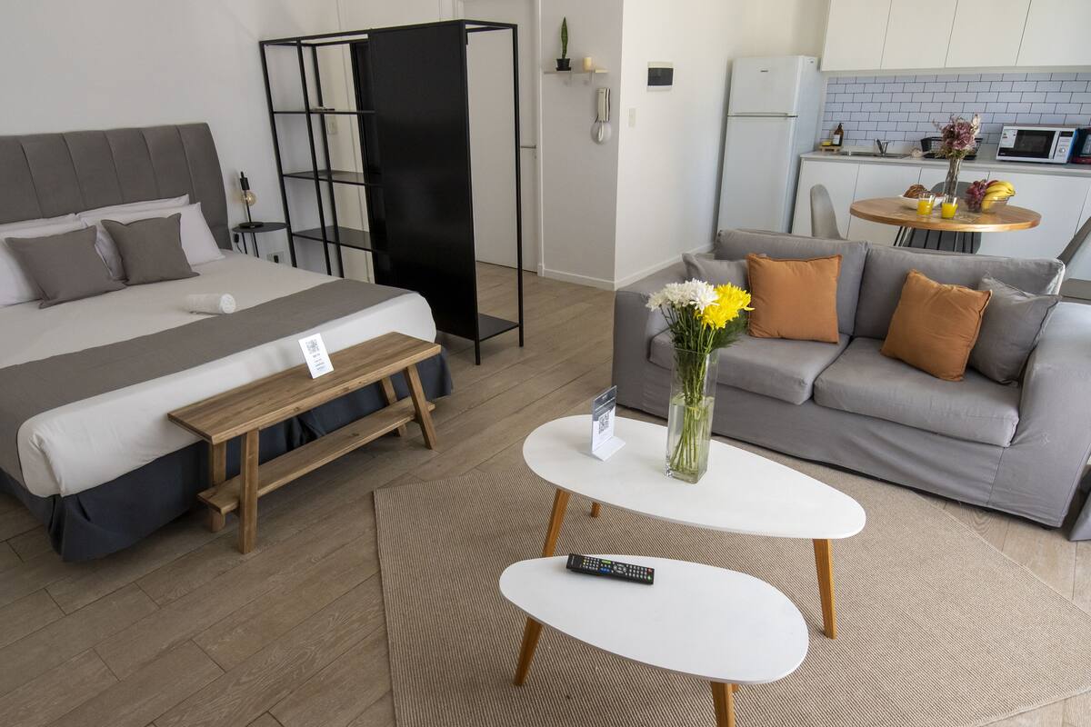 A modern hotel room with a bed, a sofa, a small table, and a kitchen area in the background.