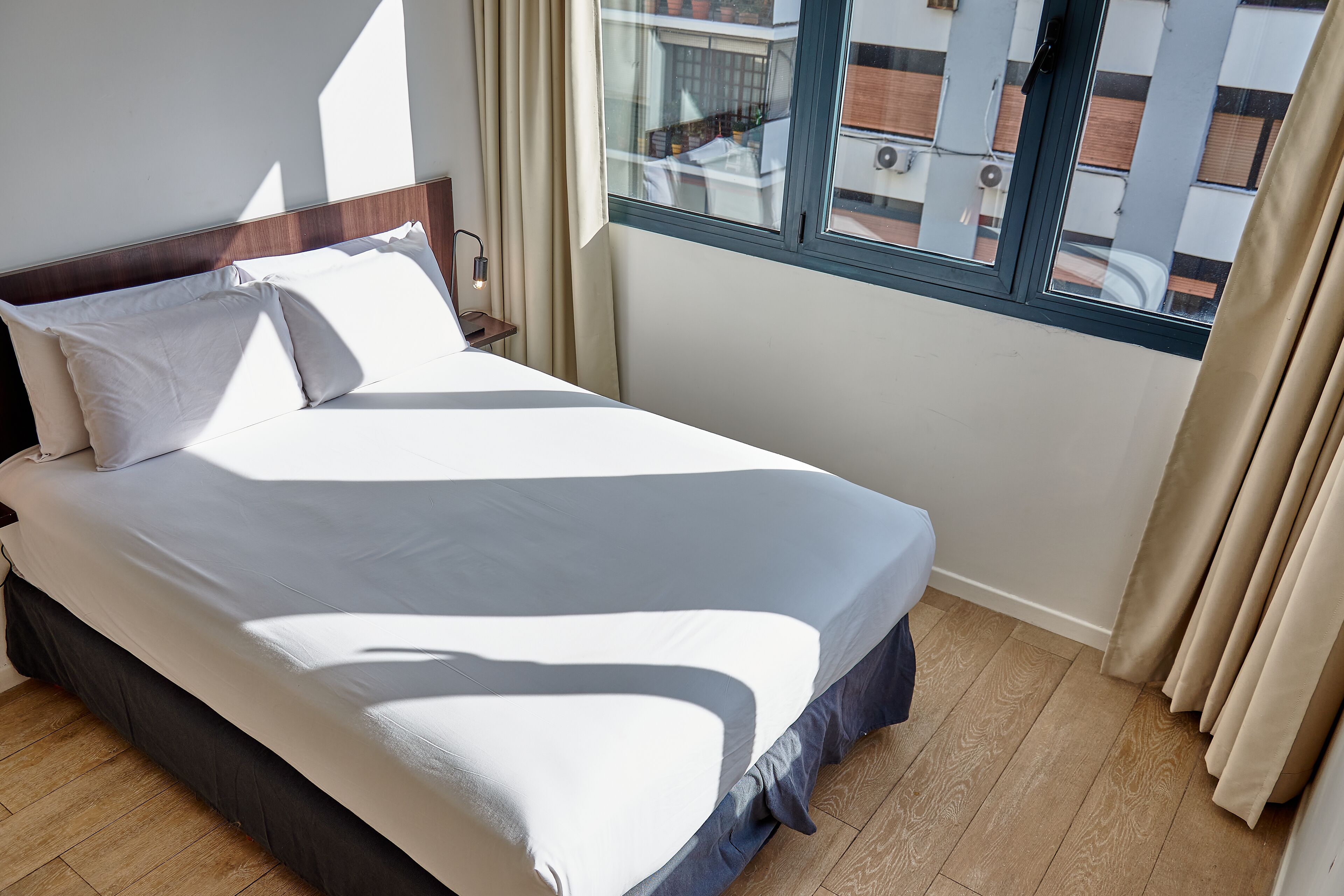 Ein ordentlich bezogenes Bett mit weißer und grauer Bettwäsche in einem Zimmer mit Holzboden und einem Vorhangfenster.