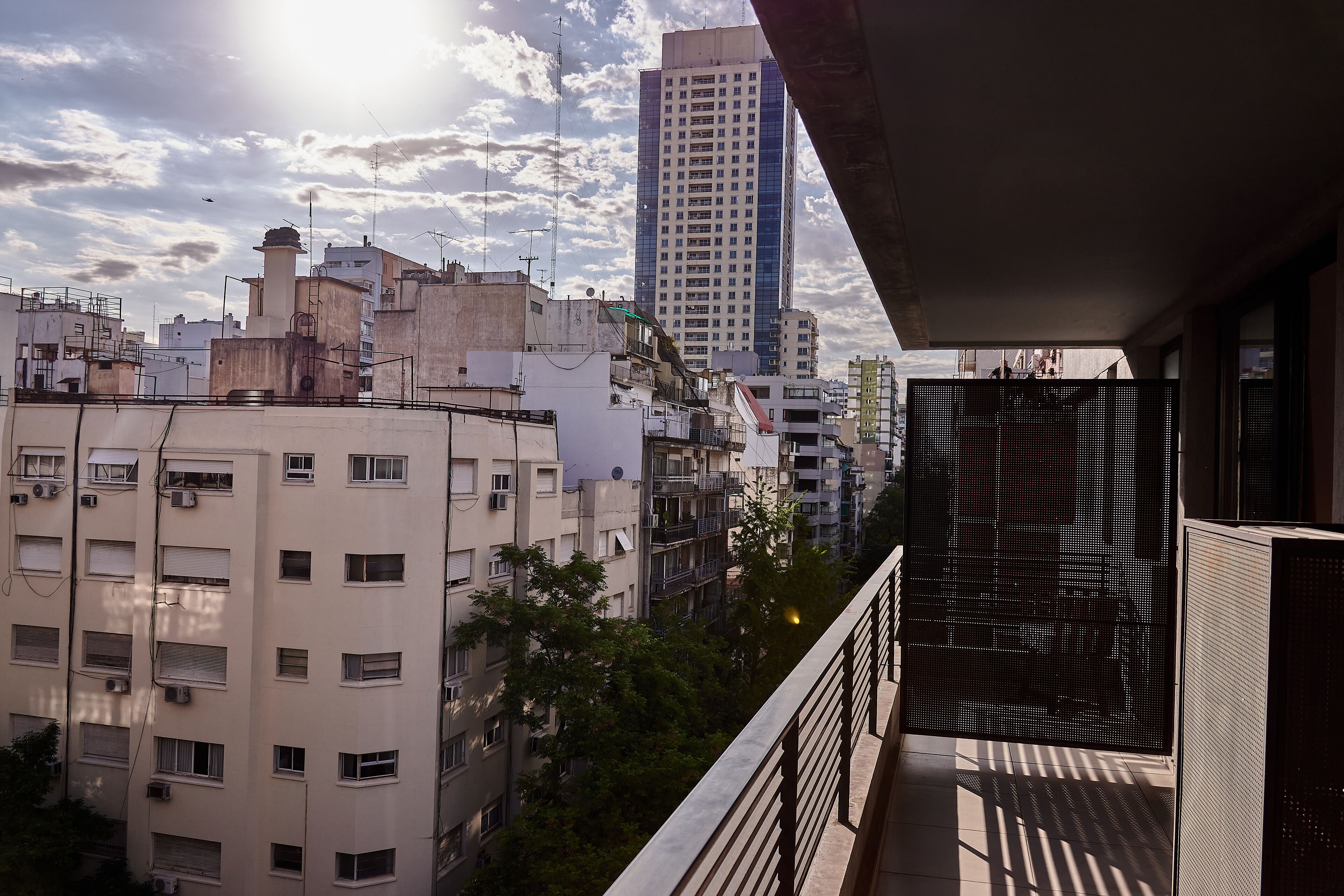 Une vue sur un paysage urbain, avec un grand immeuble en arrière-plan, un balcon doté d’une rambarde en métal et un ciel dégagé parsemé de nuages.