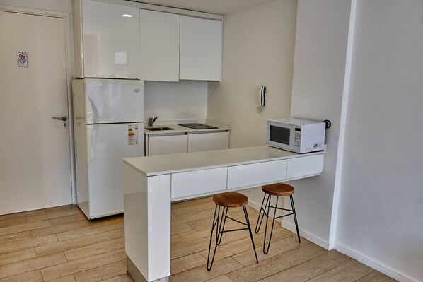 A compact kitchen with white cabinetry, a built-in microwave, a sink, and two stools.