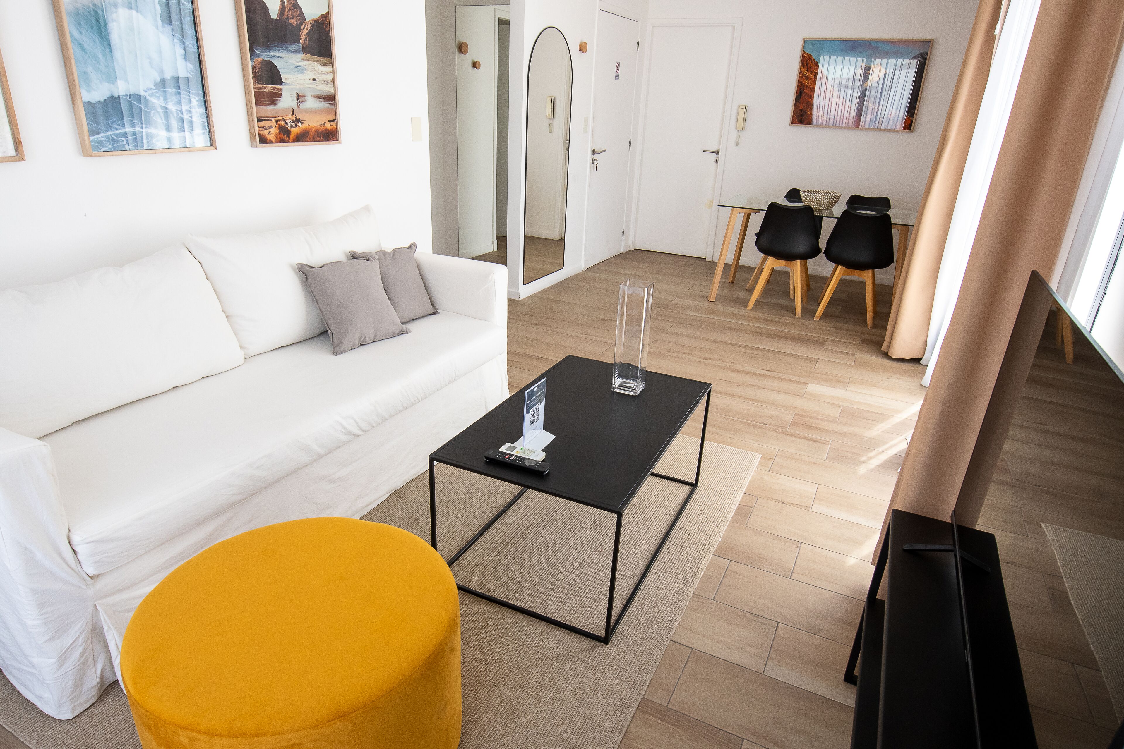 A modern living room with a white sofa, a yellow ottoman, a black coffee table, and a dining area with two chairs and a table.