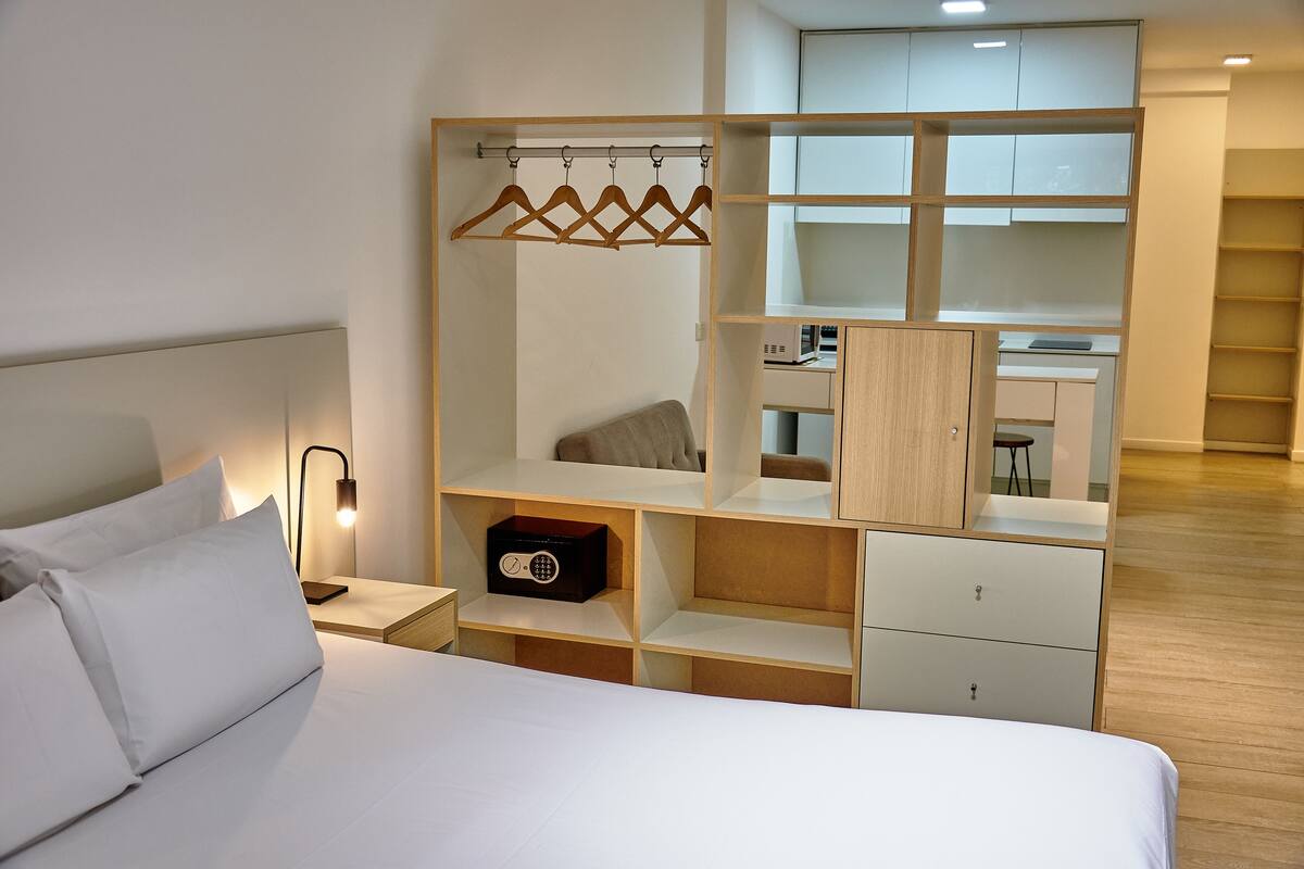 A modern hotel room with a neatly made bed, a wooden wardrobe with multiple compartments, and a small desk area with a lamp.