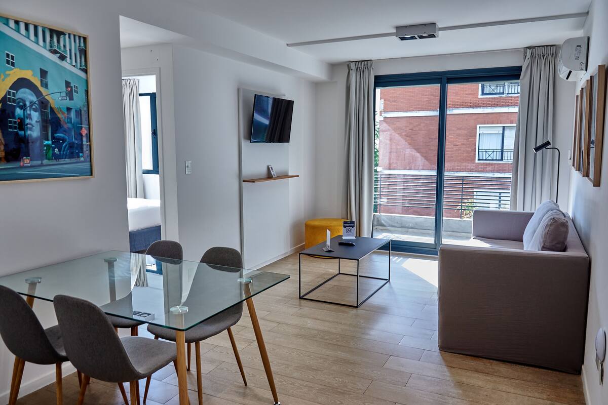 A modern living room with a glass dining table, grey chairs, a sofa, and a TV mounted on the wall.