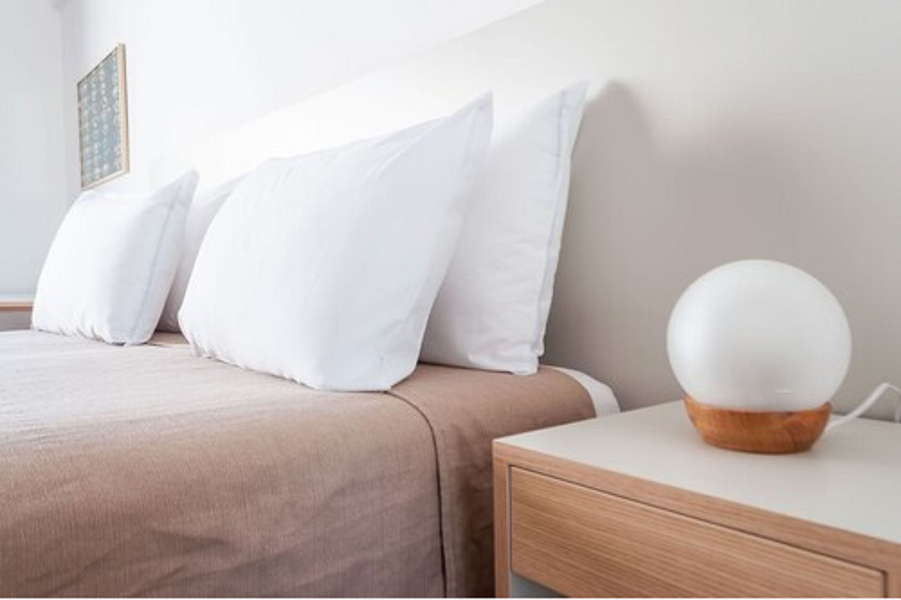 A neatly made bed with white pillows and a wooden bedside table with a white spherical object on it.