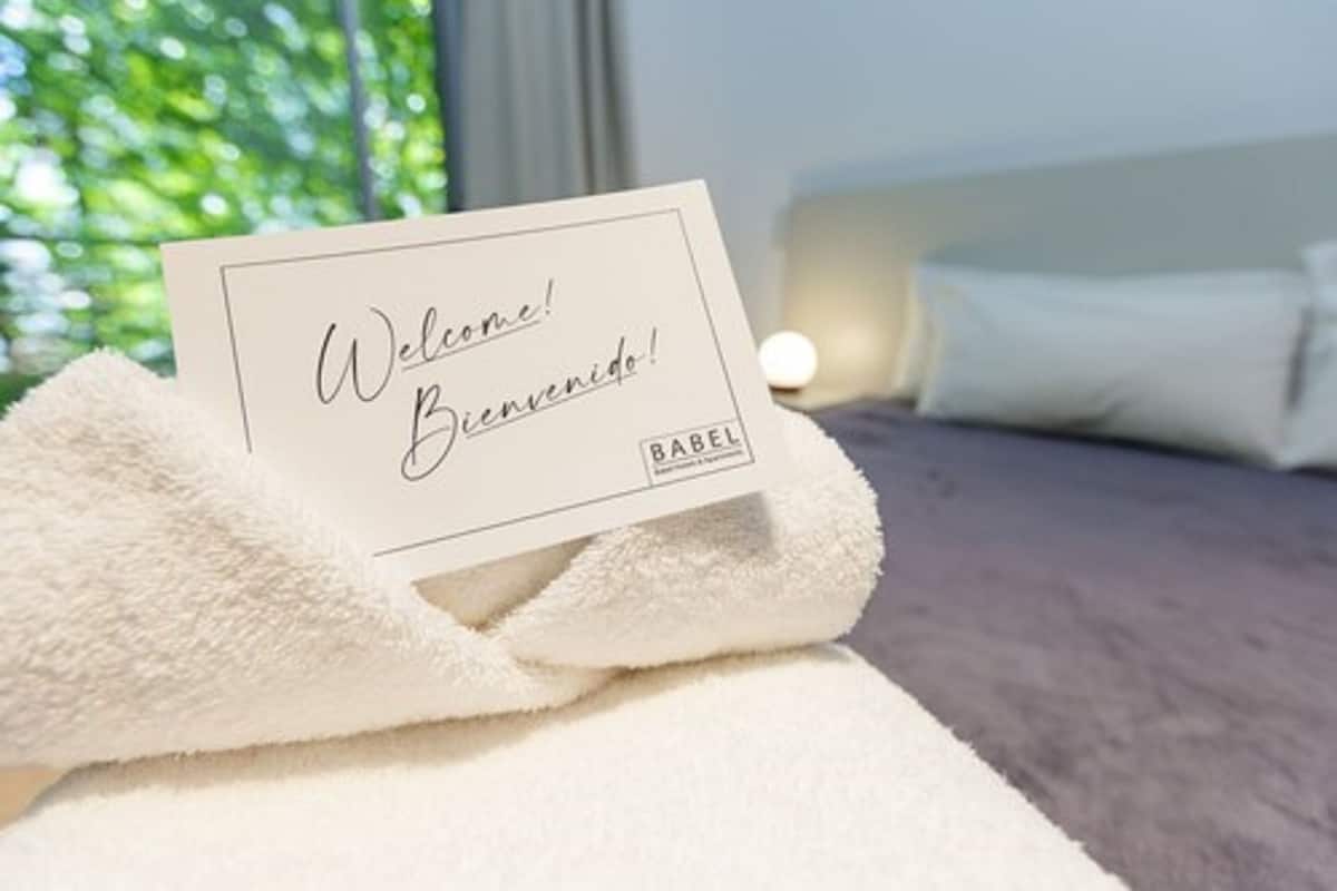 A welcome card on a white towel in a hotel room with a bed and a window with greenery outside.