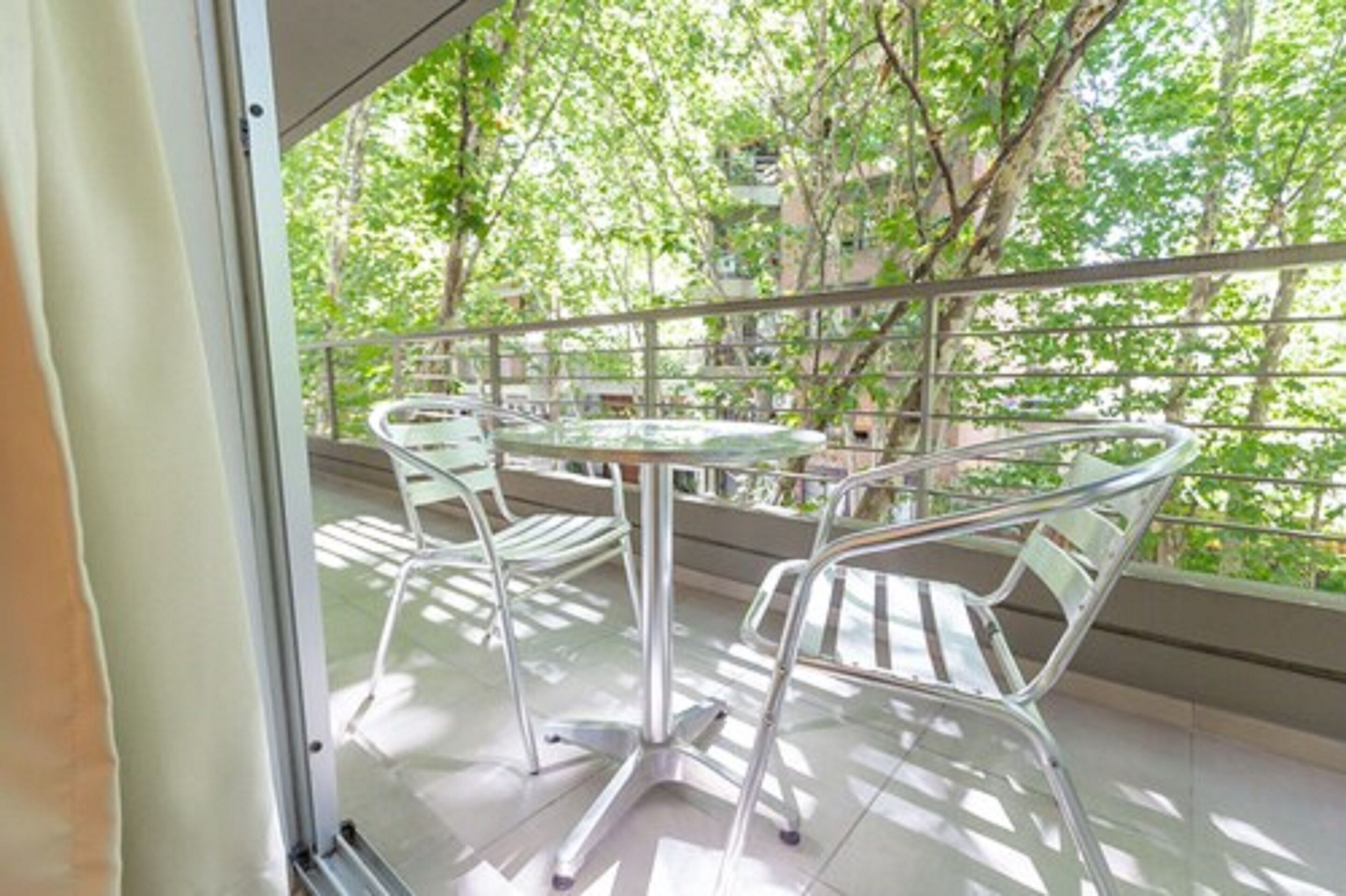 A balcony with a glass table and metal chairs, overlooking a green area with trees and buildings.