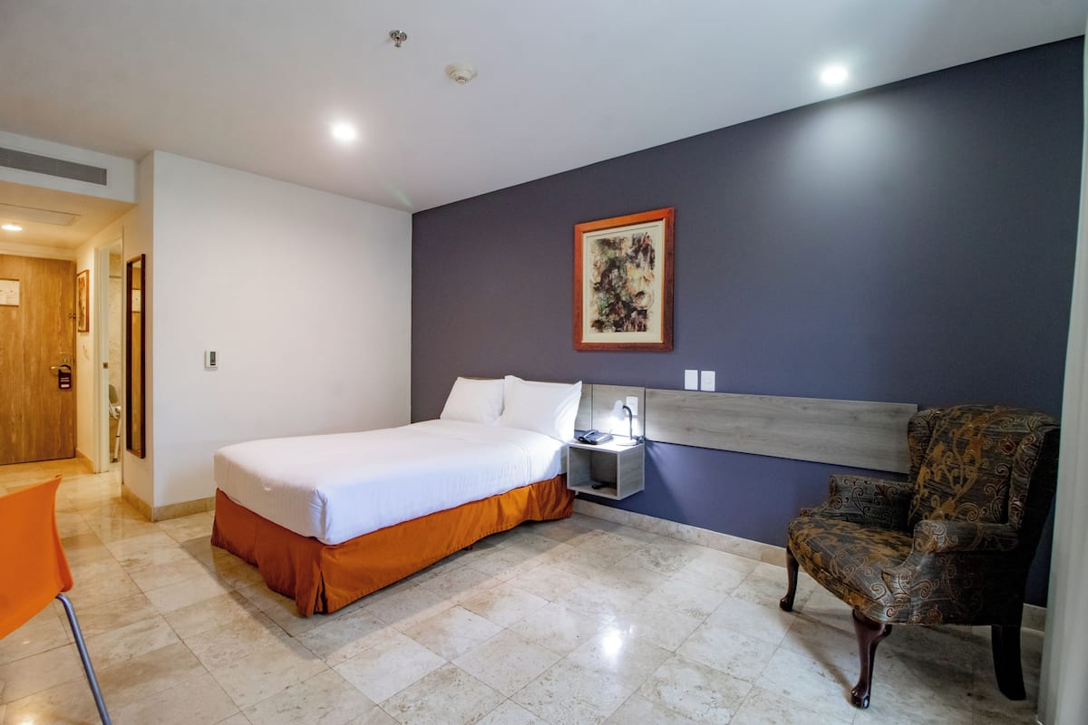 Una habitación de hotel con cama, una silla, una mesita de noche y un cuadro en la pared.