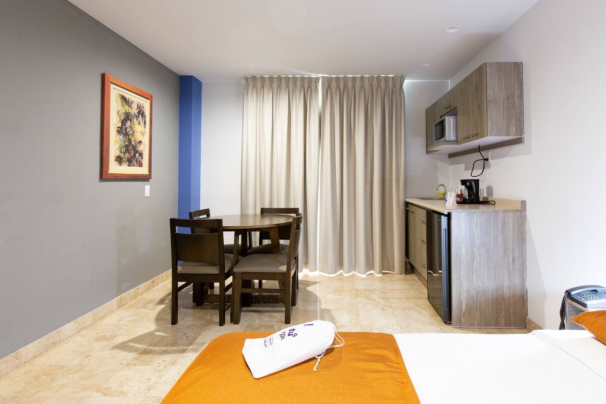 Una habitación de hotel con cama, escritorio, silla, kitchenette y cortina.