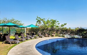 Un área junto a la piscina con sillones de descanso y sombrillas, rodeada de follaje y un cielo despejado.