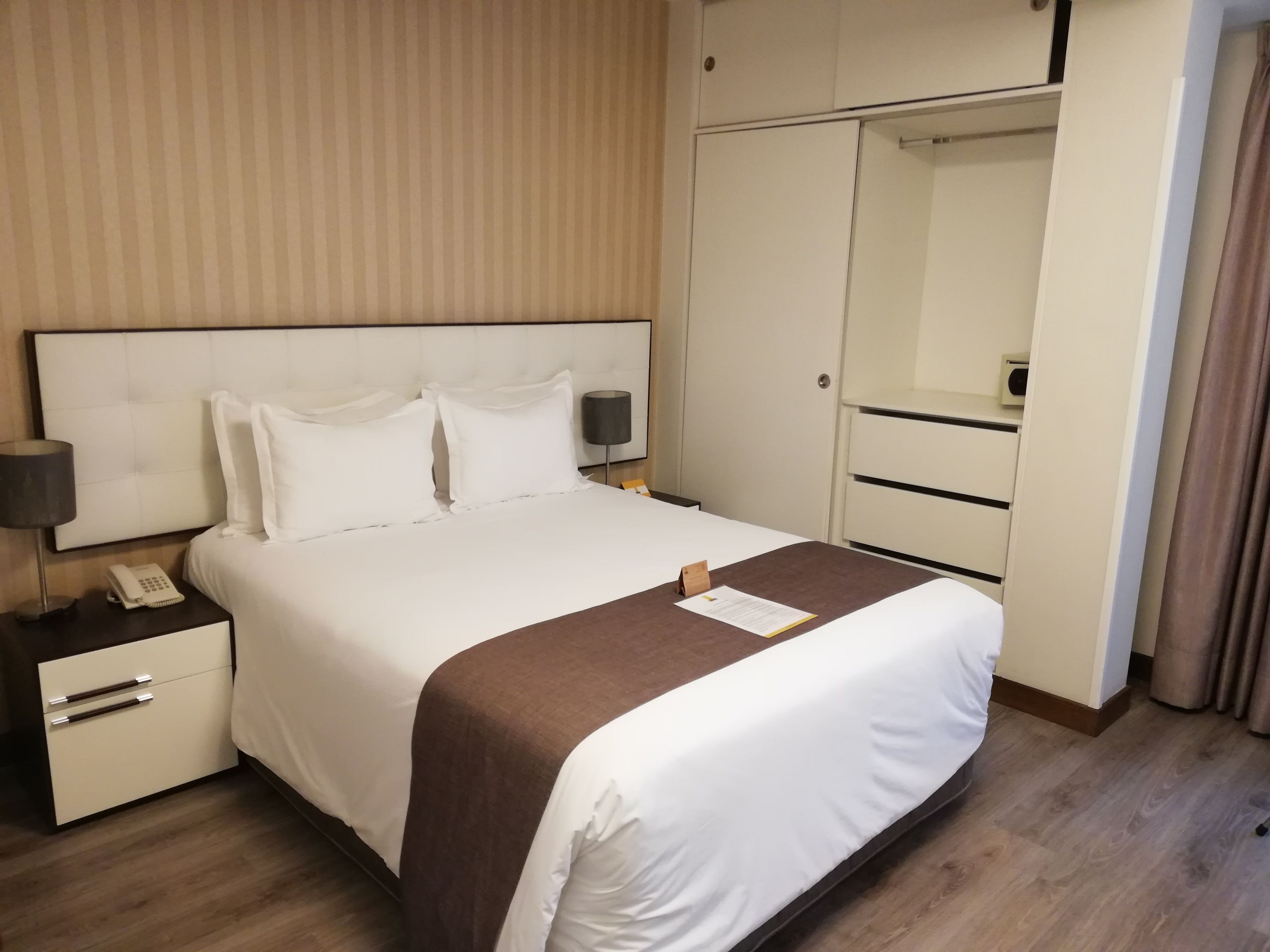 A hotel room with a large bed, a bedside table with a lamp, a telephone, and a wardrobe.