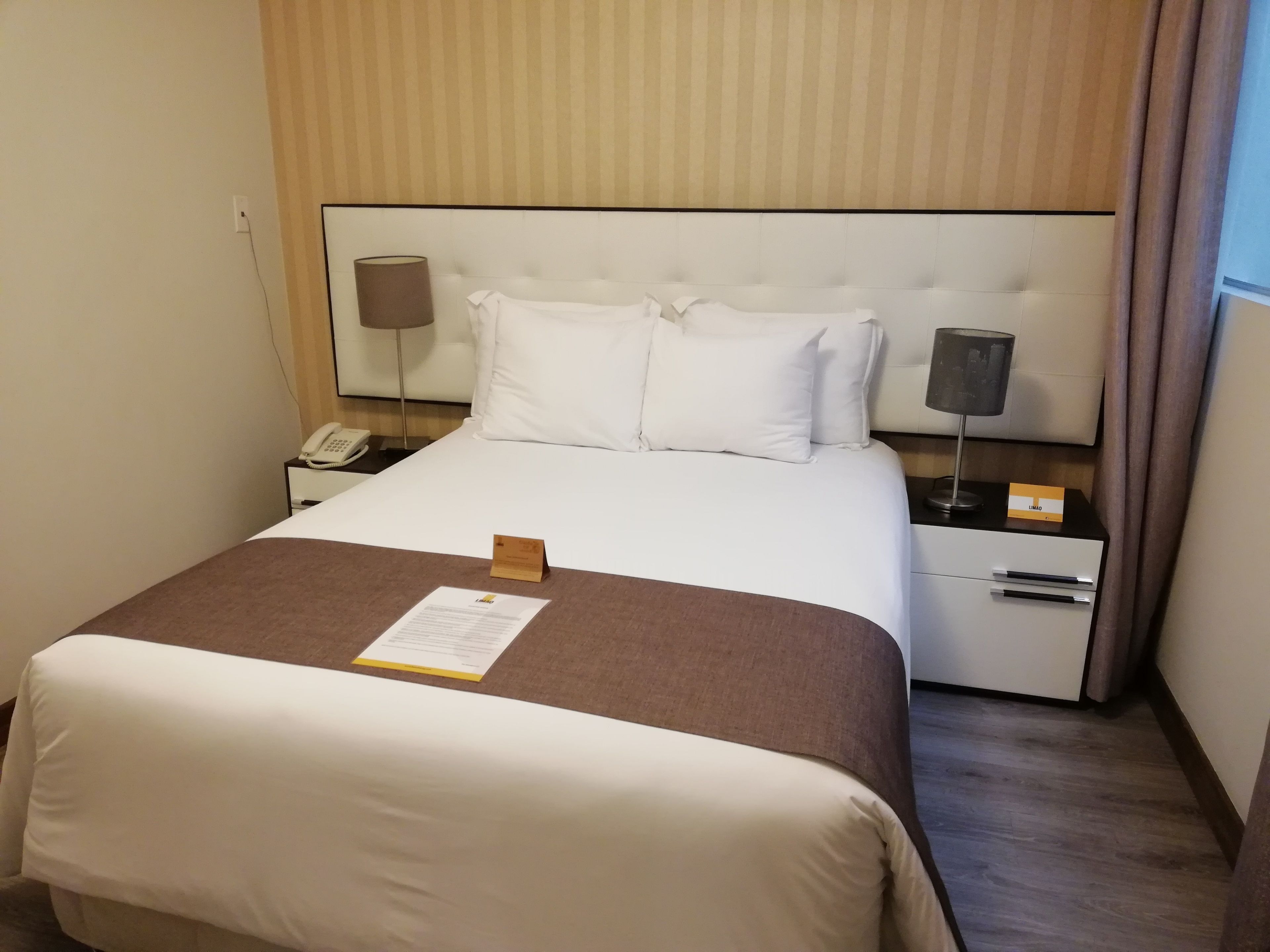 A hotel room with a neatly made bed, a bedside table with a lamp, a phone, and a small card on the table.