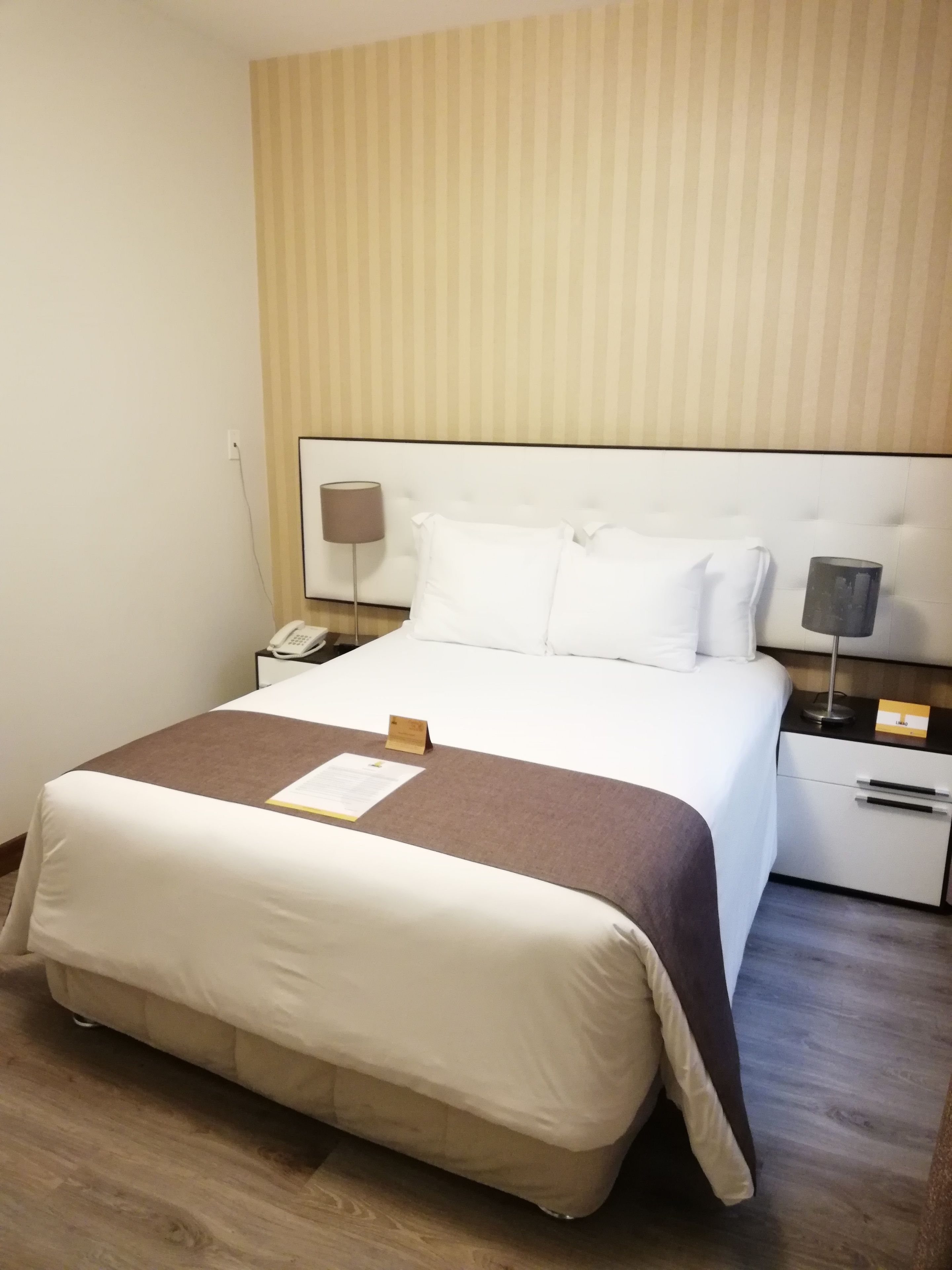 A hotel room with a large bed, a bedside table with a lamp, a phone, and a small card on the bed.