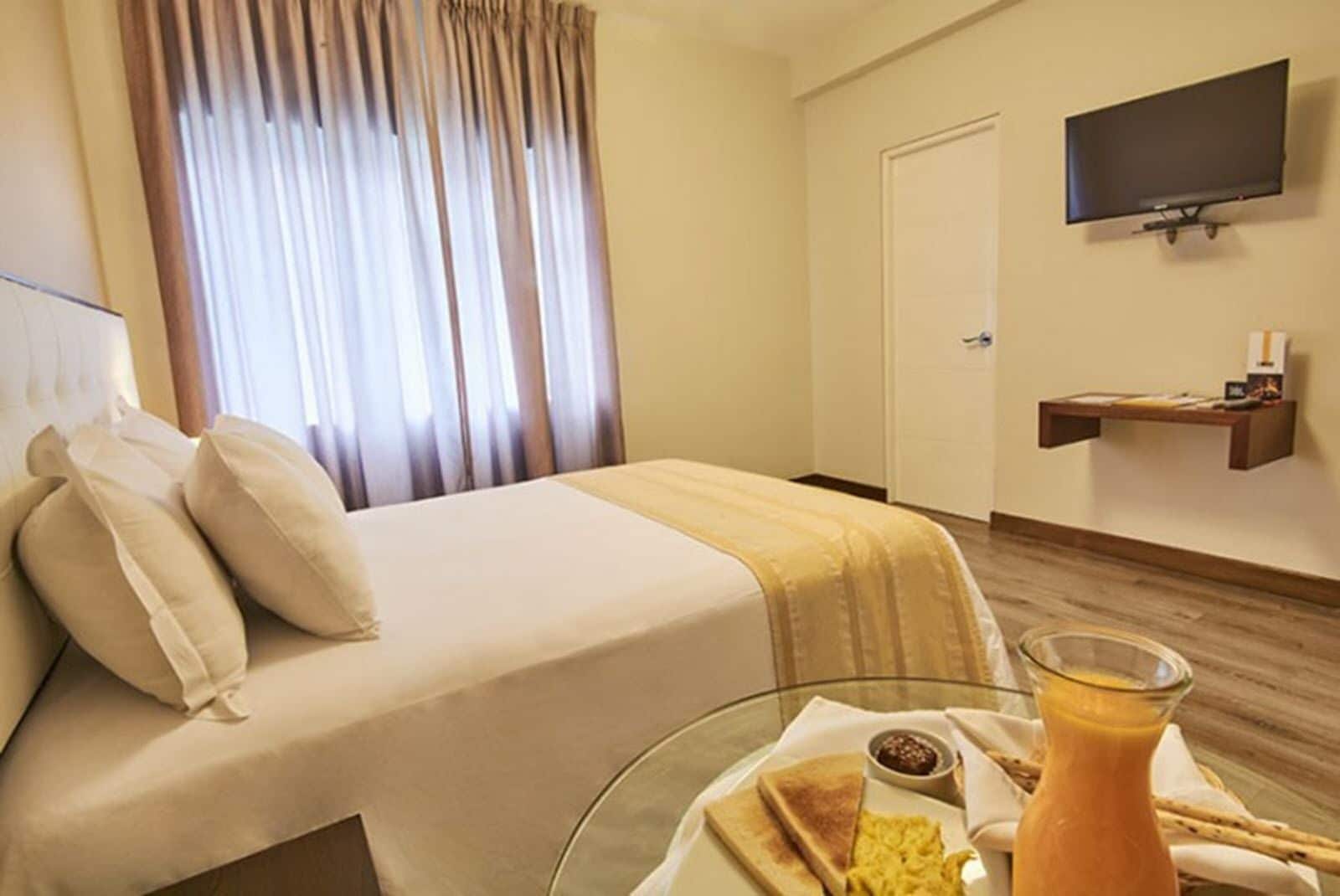 Camera d'albergo con un letto, una televisione montata a parete, un piccolo ripiano con oggetti vari e un vassoio per la colazione su un tavolino di vetro.