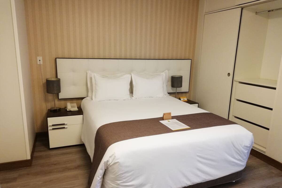Un lit bien fait, avec du linge de lit blanc, un plaid marron et une table de chevet sur laquelle se trouvent une lampe et un téléphone.