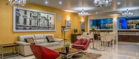 A modern hotel lobby with a sofa, red chairs, a bar area, and a large cityscape painting on the wall.