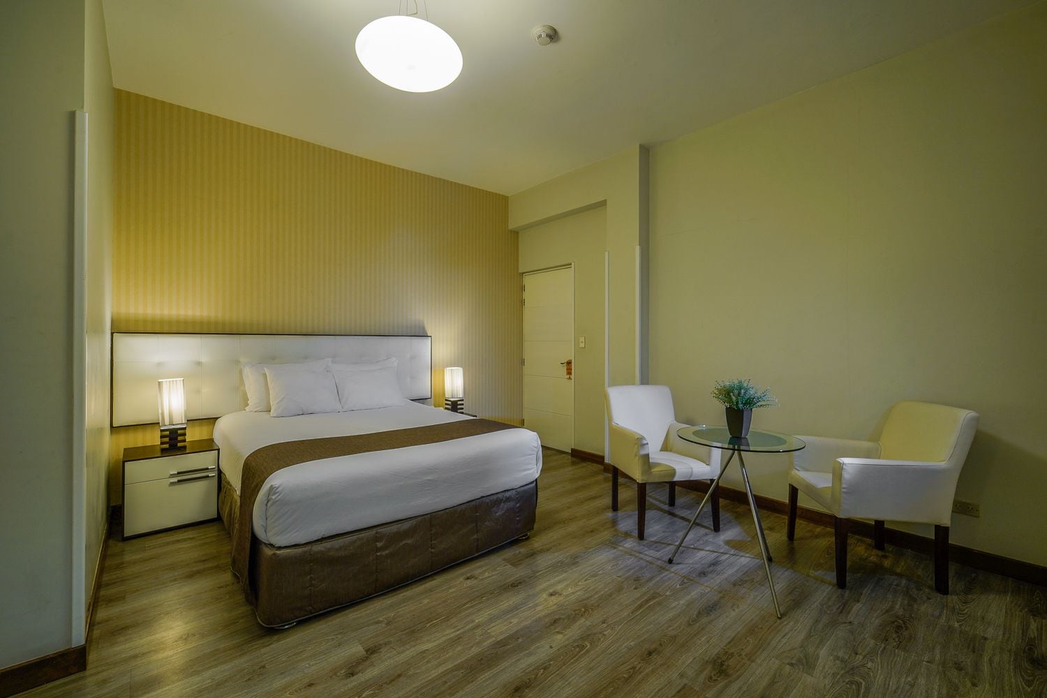Habitación de hotel con cama, mesitas de noche, una silla, una pequeña mesa con una planta y una puerta.