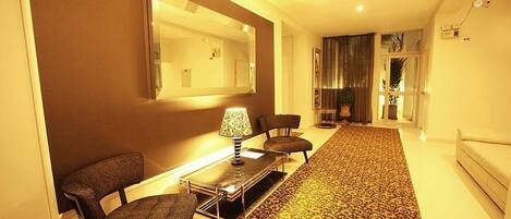 Un hall de hotel con una alfombra estampada, un sofá, una mesa con lámpara y un espejo grande en la pared.