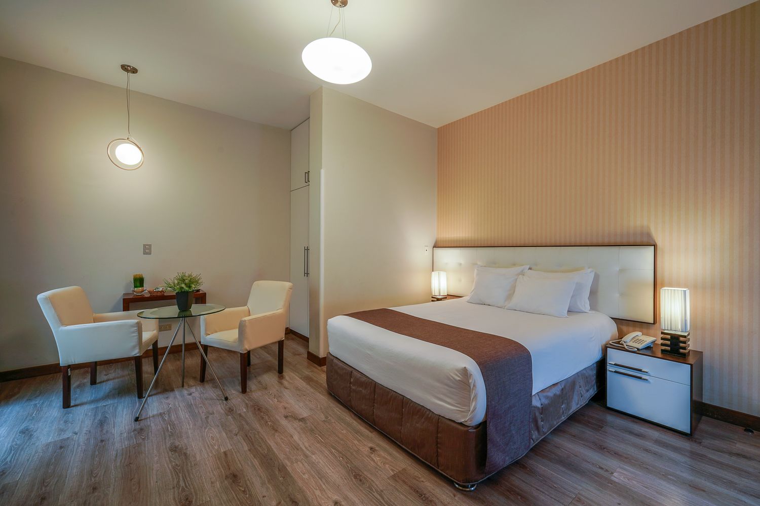 A hotel room with a bed, bedside table, lamp, chair, and a small table with a plant.