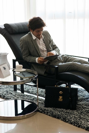 Un hombre con traje gris sentado en una silla de cuero negro, sosteniendo una tableta. Hay una mesita redonda pequeña con una jofaina blanca, una taza y un cuaderno. Un bolso de mano negro está en el suelo junto a la silla.