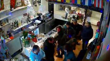 Zona de bar con estantes llenos de botellas y vasos, una barra y un área de asientos con mesas.