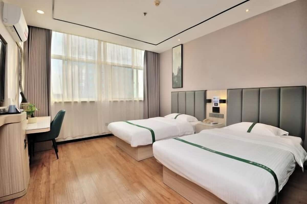A hotel room with two beds, a desk, a chair, a window with curtains, and a wall-mounted air conditioner.