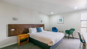 Un dormitorio con cama, mesita de noche, dos sillas, luz empotrada en la pared y un cuadro.