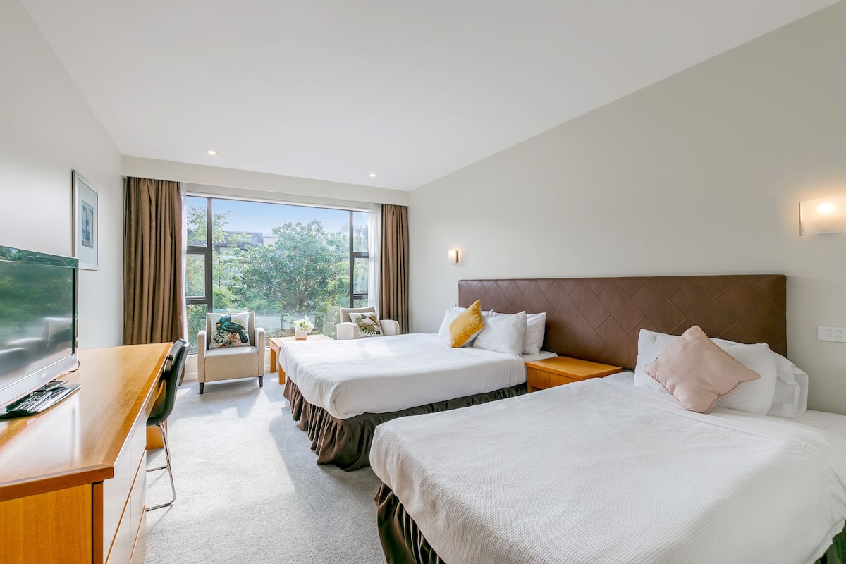 A hotel room with two beds, a desk, a chair, and a large window offering a view of trees.
