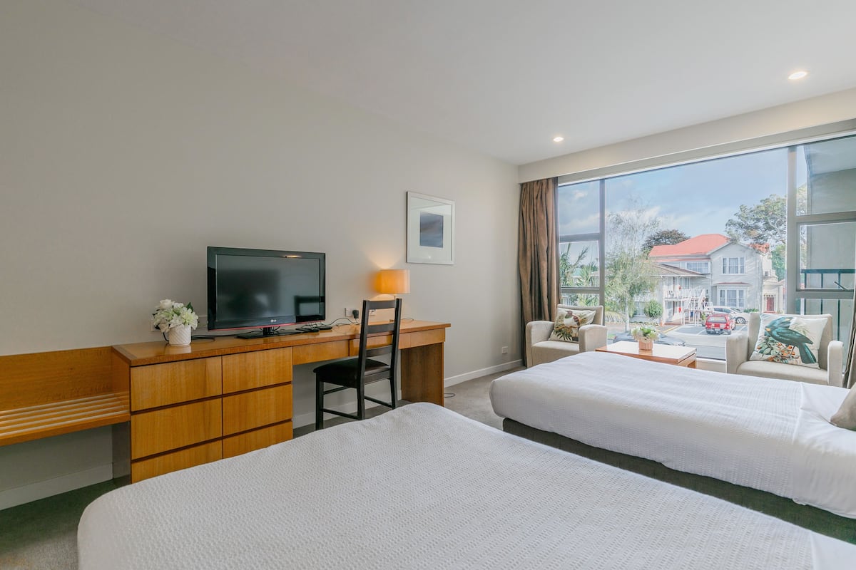 A hotel room with two beds, a desk with a TV, a chair, and a view of the street.