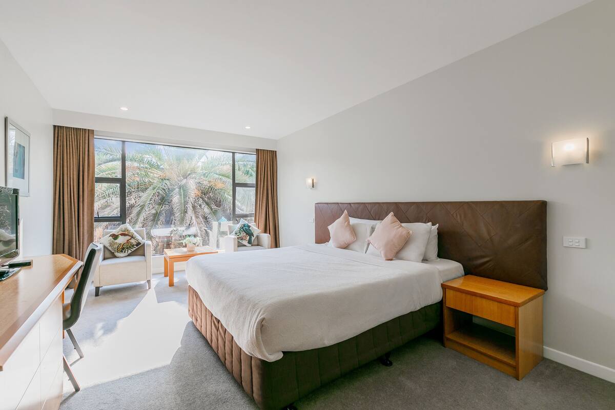 A modern hotel room with a large bed, a desk, and a view of palm trees through the window.