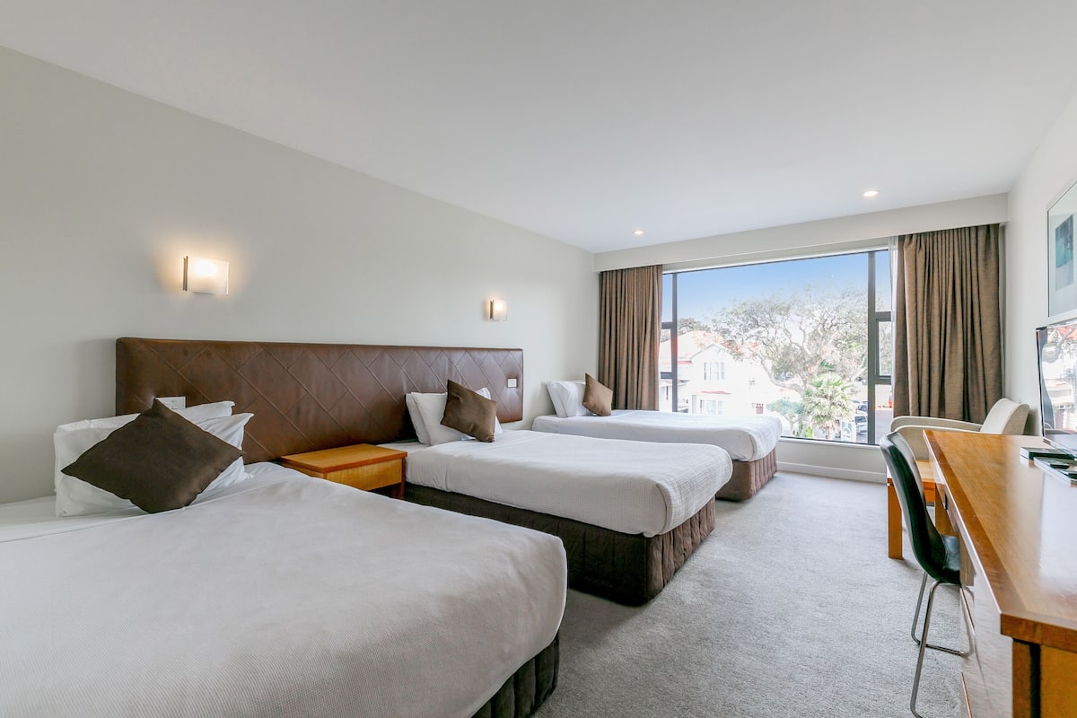 A hotel room with three beds, a wooden desk, and a large window with a view of buildings and trees.